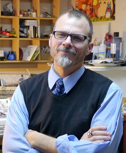 Photo. Joe Ollman, arms folded, seated in front of his drawing desk.